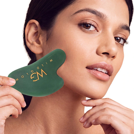 A woman holds a dark green facial gua sha massage tool with gold lettering against her cheek, shown in a close-up shot against a white background.