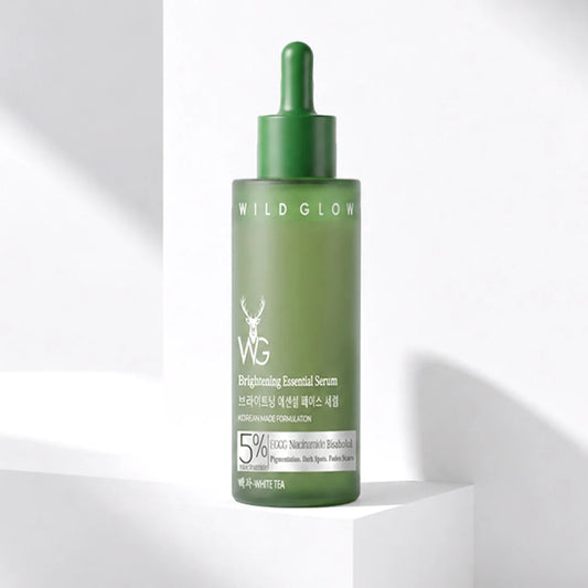 A green dropper bottle of WILDGLON Brightening Essential Serum stands on a white geometric pedestal against a white background with soft shadows.