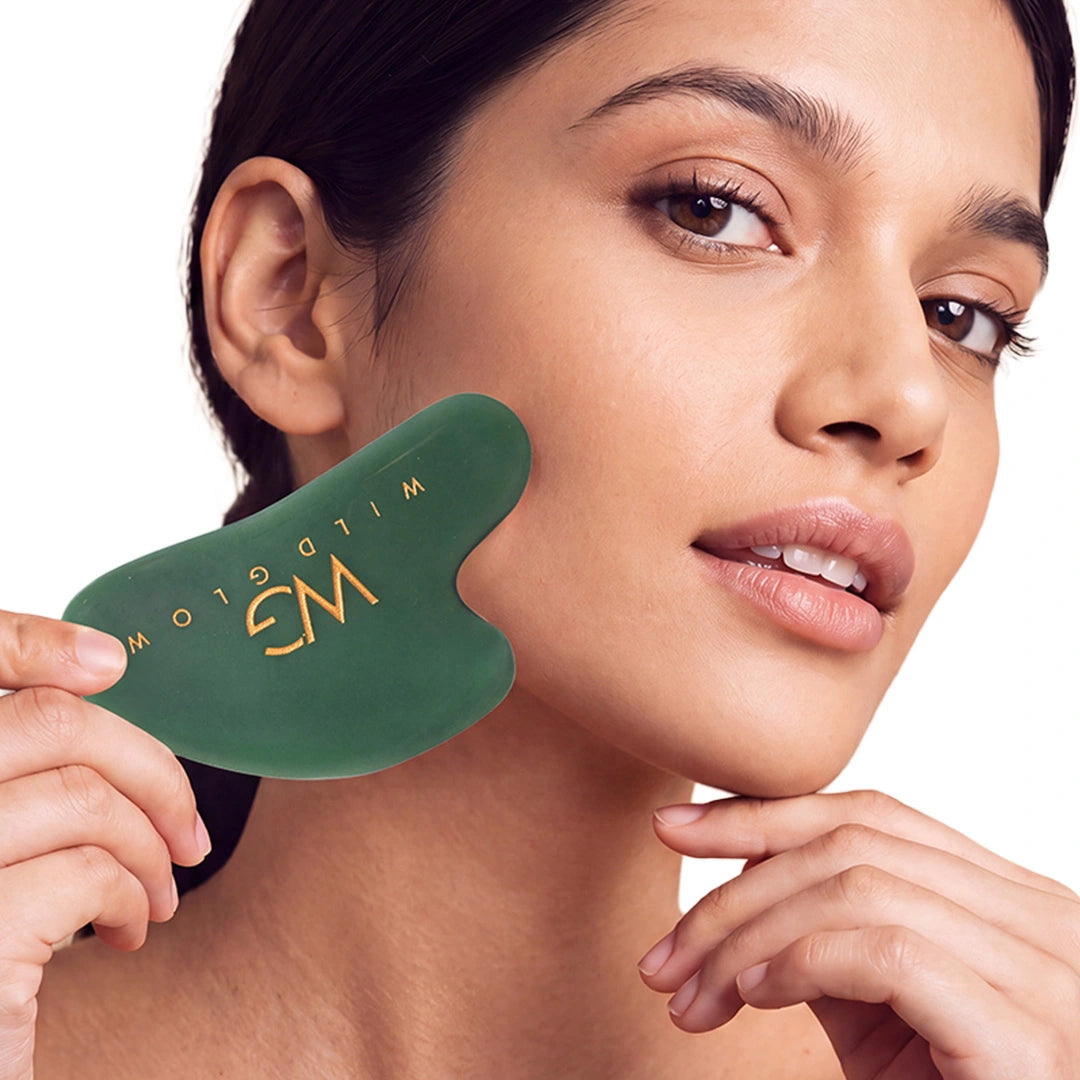 A woman holds a dark green facial gua sha massage tool with gold lettering against her cheek, shown in a close-up shot against a white background.