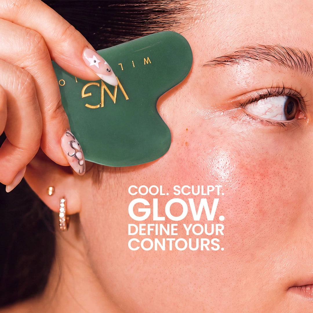 A close-up of a woman's face with a dark green facial gua sha massage tool held against her cheek. The tool has gold lettering that reads 'EM' and 'WILM'.