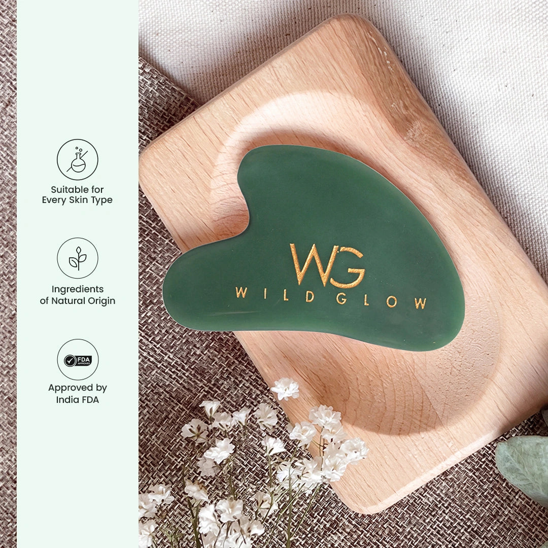 Green gua sha facial massage tool with gold 'WILD GLOW' logo, resting on a wooden tray with decorative flowers and a light green informational sidebar on the left.