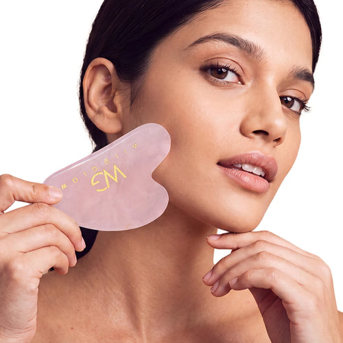 A woman holding a translucent pink gua sha facial massage tool against her neck, with gold lettering on the tool, on a solid white background.