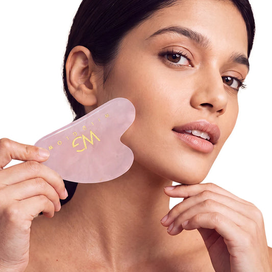 A woman holding a translucent pink gua sha facial massage tool against her neck, with gold lettering on the tool, on a solid white background.