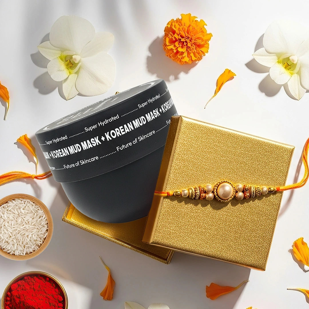 A black jar of Korean mud mask next to a gold gift box with a gold and orange rakhi, styled with white flowers, marigold, rice, red powder, and orange petals on a white background.